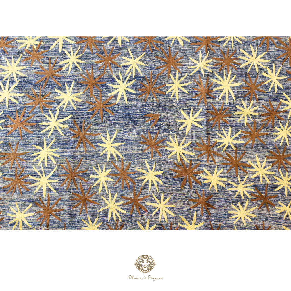 Patterned rug with starburst design in brown and yellow on a blue background, featuring a brand logo at the bottom.