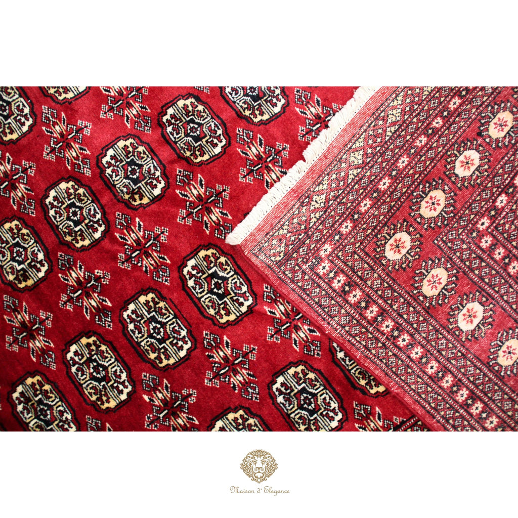 Close-up of a red patterned fabric with intricate designs, featuring a brand logo at the bottom.