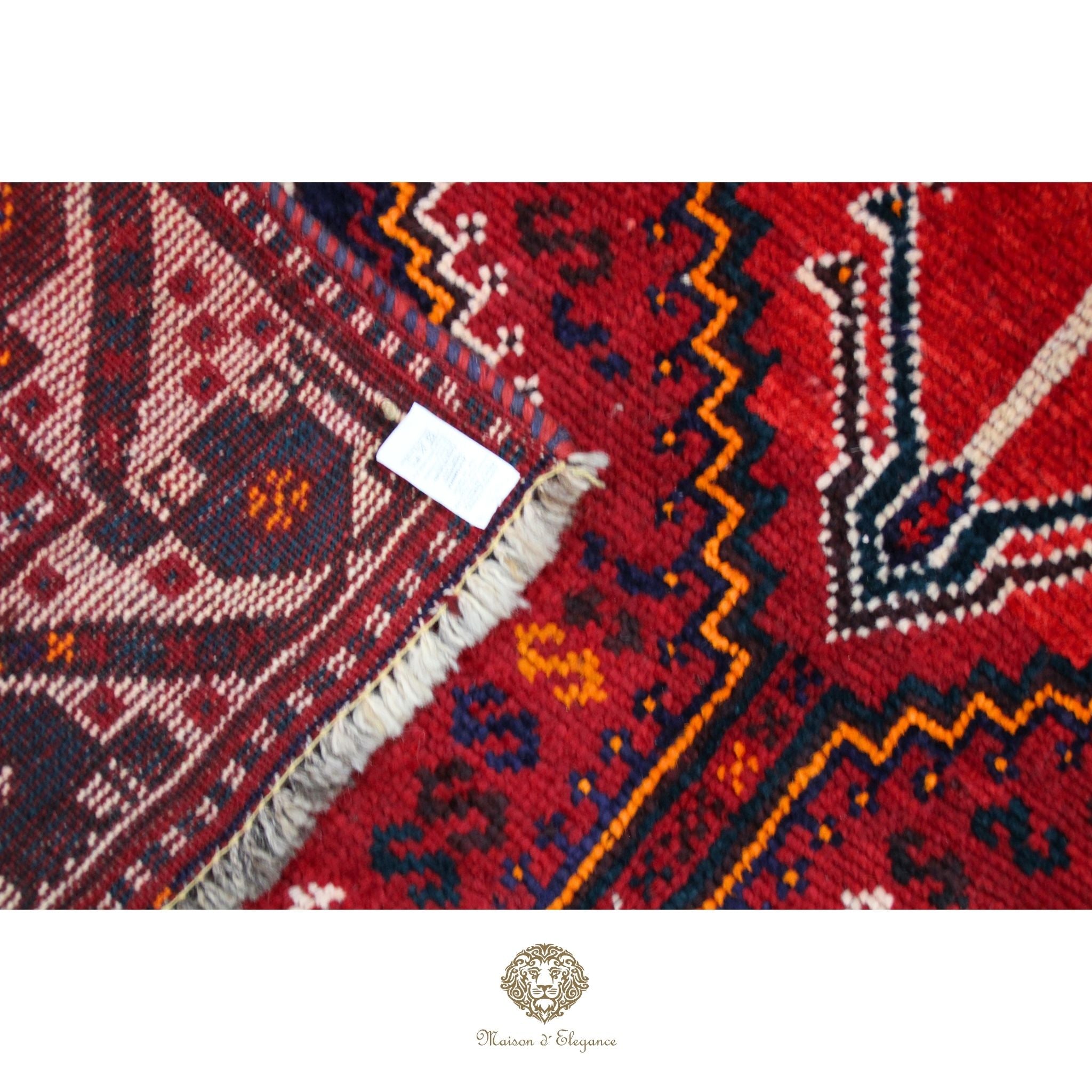 Close-up of a red patterned rug with a white label, featuring a brand logo at the bottom.