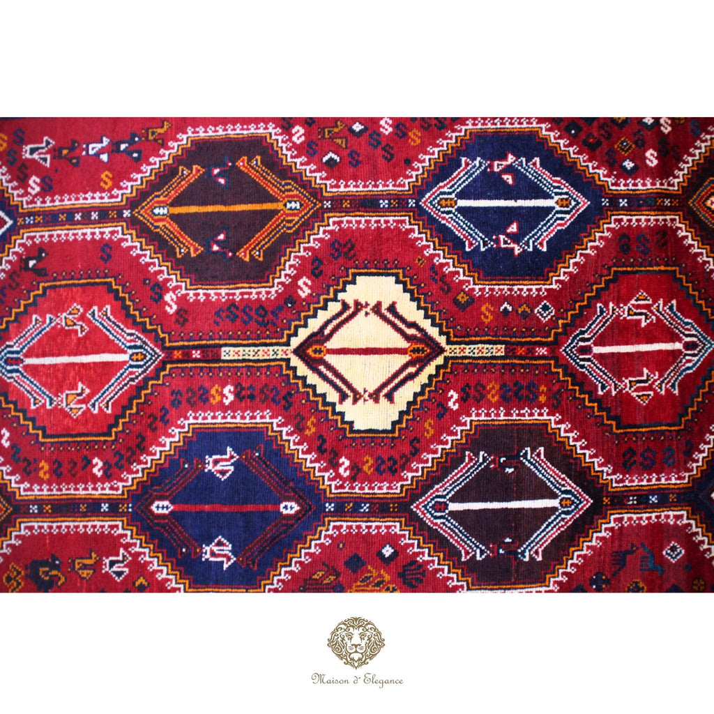 Decorative rug with geometric patterns on a red background, featuring a brand logo at the bottom.